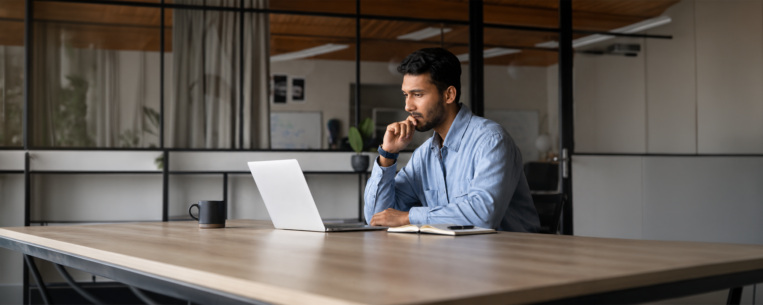 Man in a blue shirt sitting at a wooden desk, looking thoughtfully at a laptop screen with his hand resting near his chin.