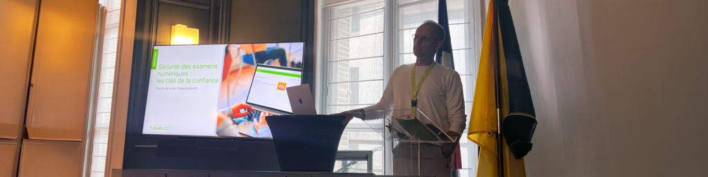A man stands at a glass podium giving a presentation. Behind him is a large screen displaying a slide with French text and an image of a laptop, alongside an open laptop on a stand. Two flags are visible in the background.