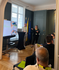 Two presenters standing at a high table in an elegant meeting room, addressing a small seated audience. A large screen displays a presentation behind them, with a laptop open on the table. The European Union and Belgian flags are visible in the background.