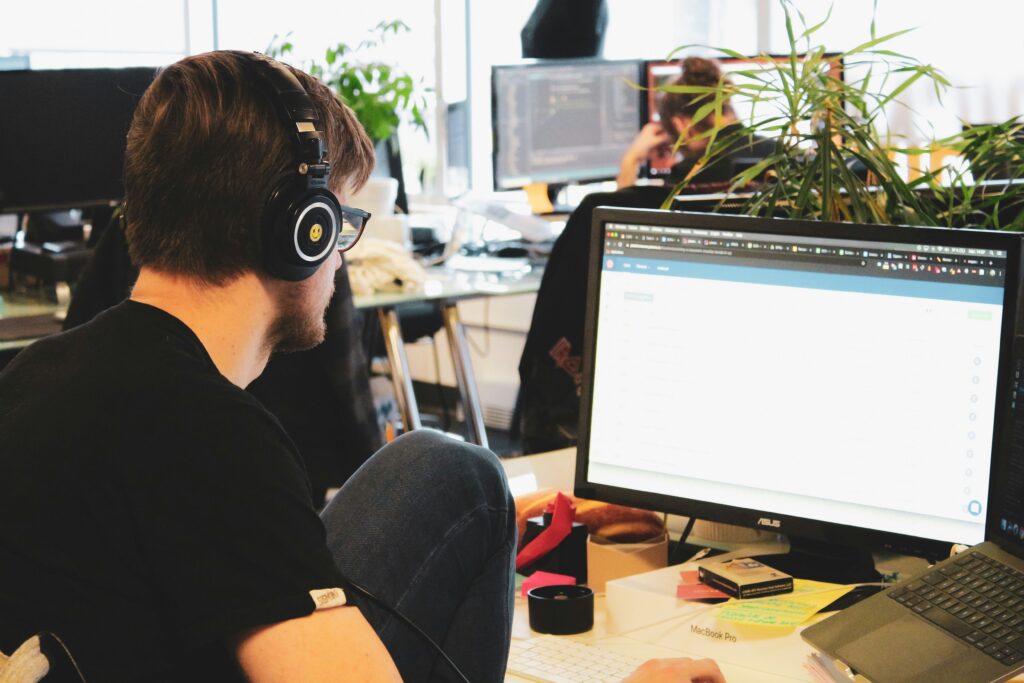 A person wearing headphones sits at a desk working on a desktop computer in a busy open-plan office. Plants and colleagues are visible in the blurred background, suggesting a focused work environment.