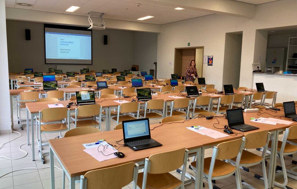 A large, mostly empty examination hall with rows of wooden desks each equipped with a laptop, ready for a computer-based assessment. A projected screen is visible at the front, and one person stands near the back of the room, suggesting the space is being prepared for an upcoming exam or test session.