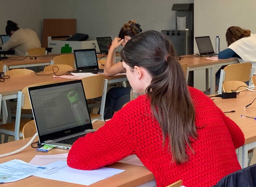 A rear view of a young woman in a red knitted jumper working on a laptop in a university examination room, with printed papers on the desk beside her. Other students with laptops are visible in the background, suggesting an in-person digital assessment or computer-based exam setting.