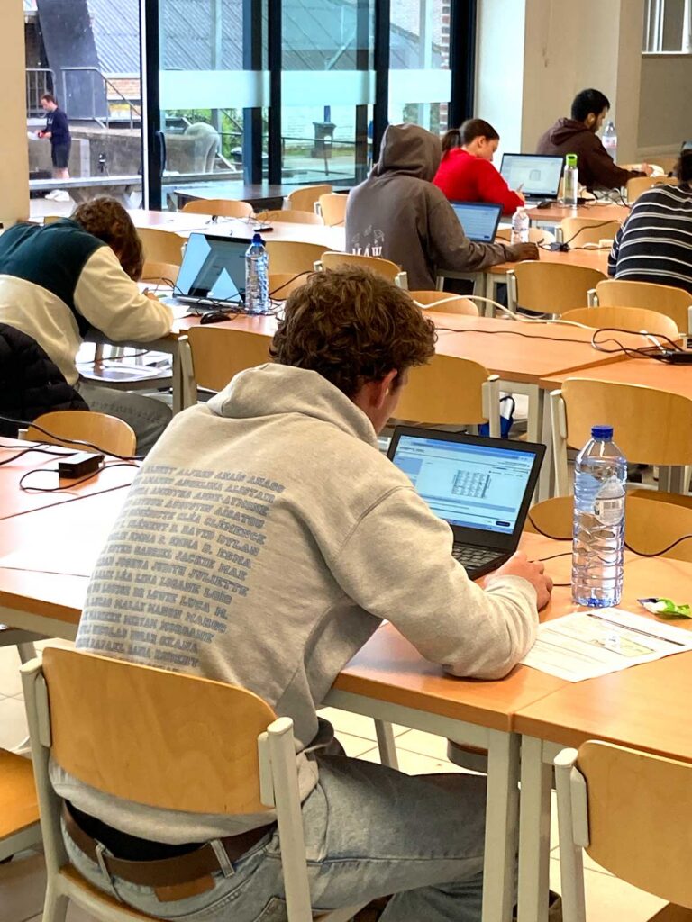 A rear view of students seated at individual wooden desks with laptops and water bottles in a bright, modern examination room with large windows. The student in the foreground is wearing a grey hoodie, conveying a focused computer-based exam environment.