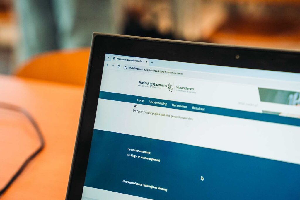 A close-up of a laptop screen displaying the website of Toelatingsexamens Vlaanderen (Flemish entrance examinations), with navigation menu items including Home, Voorbereiding, Het examen, and Resultaat. An error message indicating the requested page could not be found is visible on screen.