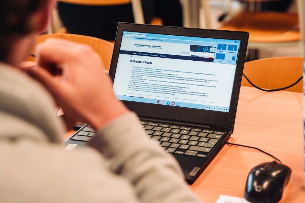 **ALT text:**

A close-up over the shoulder of a person reading content on a laptop displaying the Toelatingsexamens Vlaanderen website. A computer mouse is visible on the desk beside the laptop, suggesting a candidate reviewing exam instructions or information.