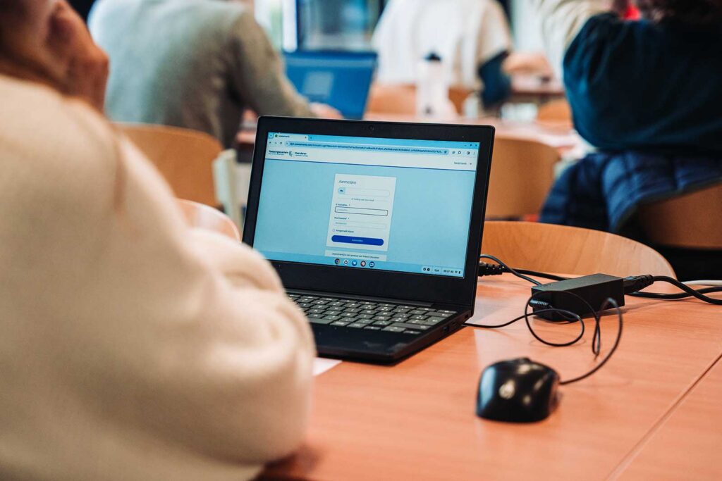 **ALT text:**

A close-up of a laptop on a wooden desk displaying a login or form page, with a computer mouse and a portable device beside it. Other students are visible in the blurred background, suggesting a computer-based examination or registration setting.