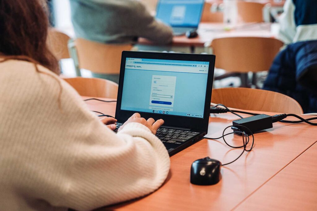 **ALT text:**

A close-up of a woman's hands typing on a laptop displaying a login or form page, with a computer mouse and a portable device on the wooden desk beside it. Other students with laptops are visible in the blurred background, suggesting a computer-based examination setting.