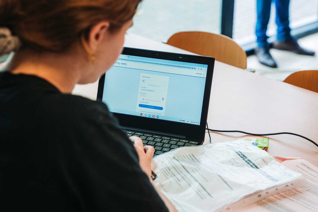 **ALT text:**

A rear view of a woman with red hair working on a laptop displaying a login or form page, with printed documents open beside the keyboard. A person's legs are visible standing in the background near a window, suggesting an invigilated computer-based examination setting.