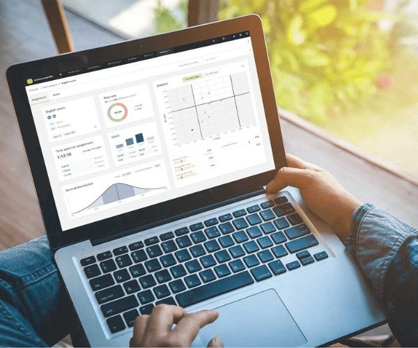 A person holding a laptop displaying the assessmentQ analytics dashboard for an English exam, showing a pass rate donut chart, score bar chart, time spent, normal distribution curve, and a statistics scatter plot, with greenery visible through a window in the background.