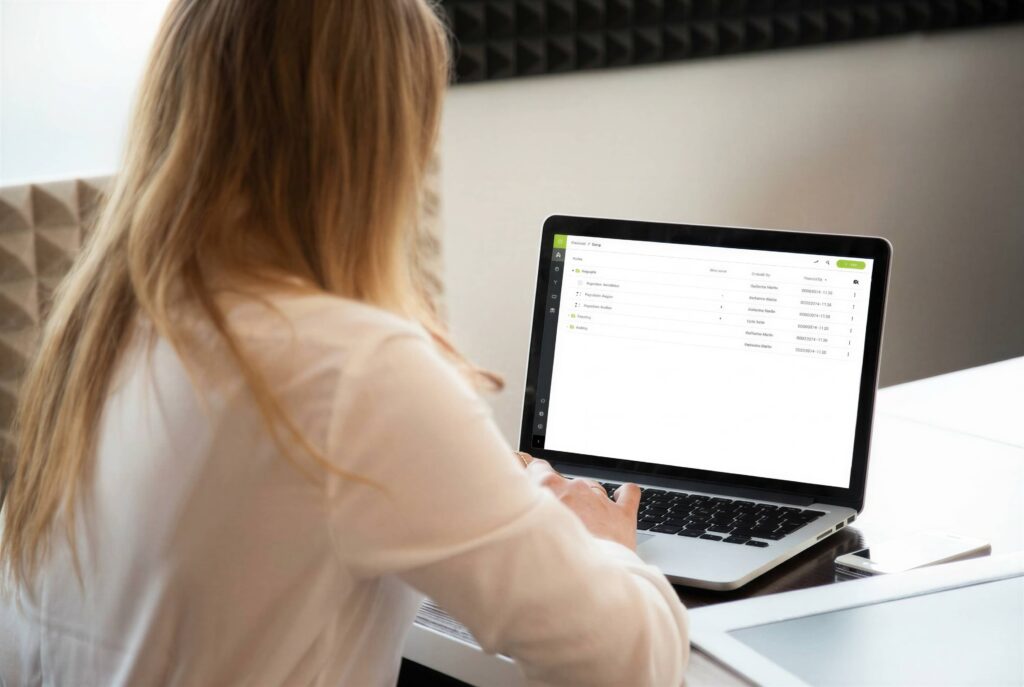 **ALT text:** A rear view of a woman with long blonde hair working on a laptop displaying a green-accented data management or assessment platform interface. She is seated at a desk in a bright, modern workspace.