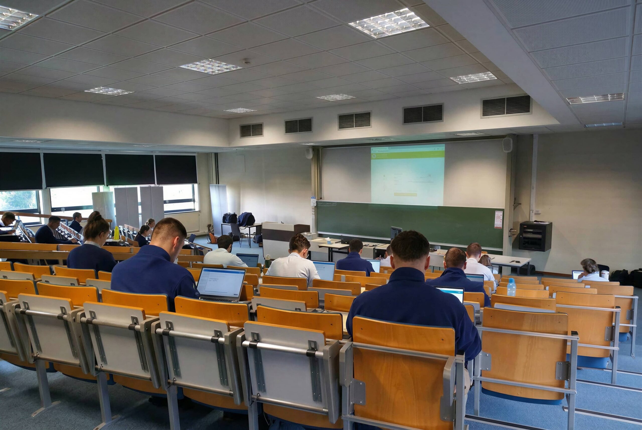 A university lecture hall with students in blue uniforms seated at wooden tiered seating, working on laptops and facing a projected green screen at the front of the room beside a blackboard, suggesting a computer-based examination or training session.