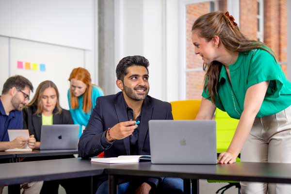 A man in a dark blazer smiles and gestures with a pen while talking with a woman in a green top who leans over a table with a laptop. Other students work in the background in a bright, modern classroom or collaborative study space with colourful furniture and sticky notes on the wall.