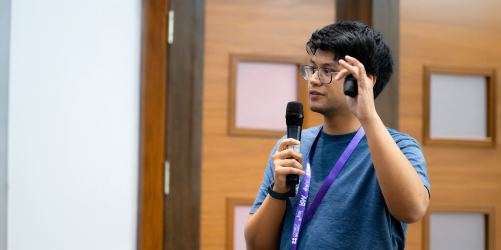 A person wearing glasses and a blue shirt speaks into a handheld microphone while giving a presentation at a conference, gesturing with a presentation remote. A purple event lanyard hangs around their neck, and the background features warm wood paneling and a projection screen area.