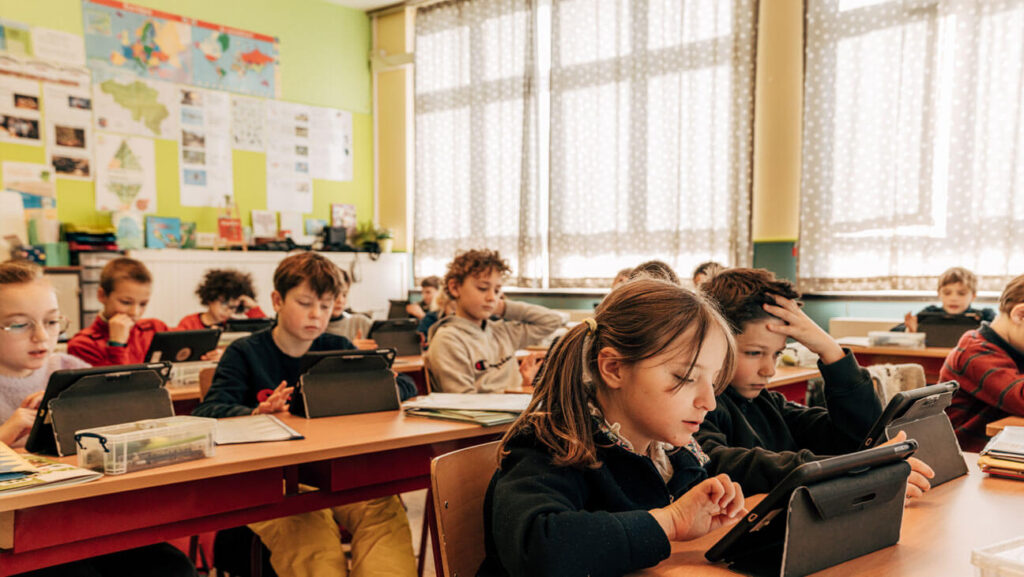 A primary school classroom with rows of children seated at wooden desks, each working on a tablet. The bright room features large curtained windows, colourful wall displays including a world map, and green walls, conveying an engaged digital learning environment.