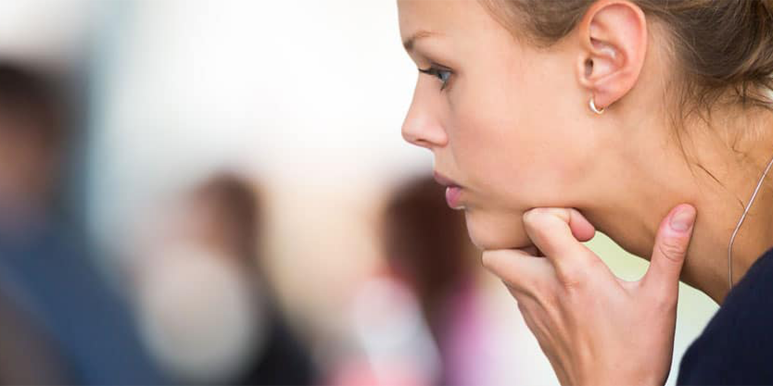 A close-up side profile of a woman resting her hand on her chin in a thoughtful pose, with blurred figures visible in the soft background, suggesting a professional or training environment.